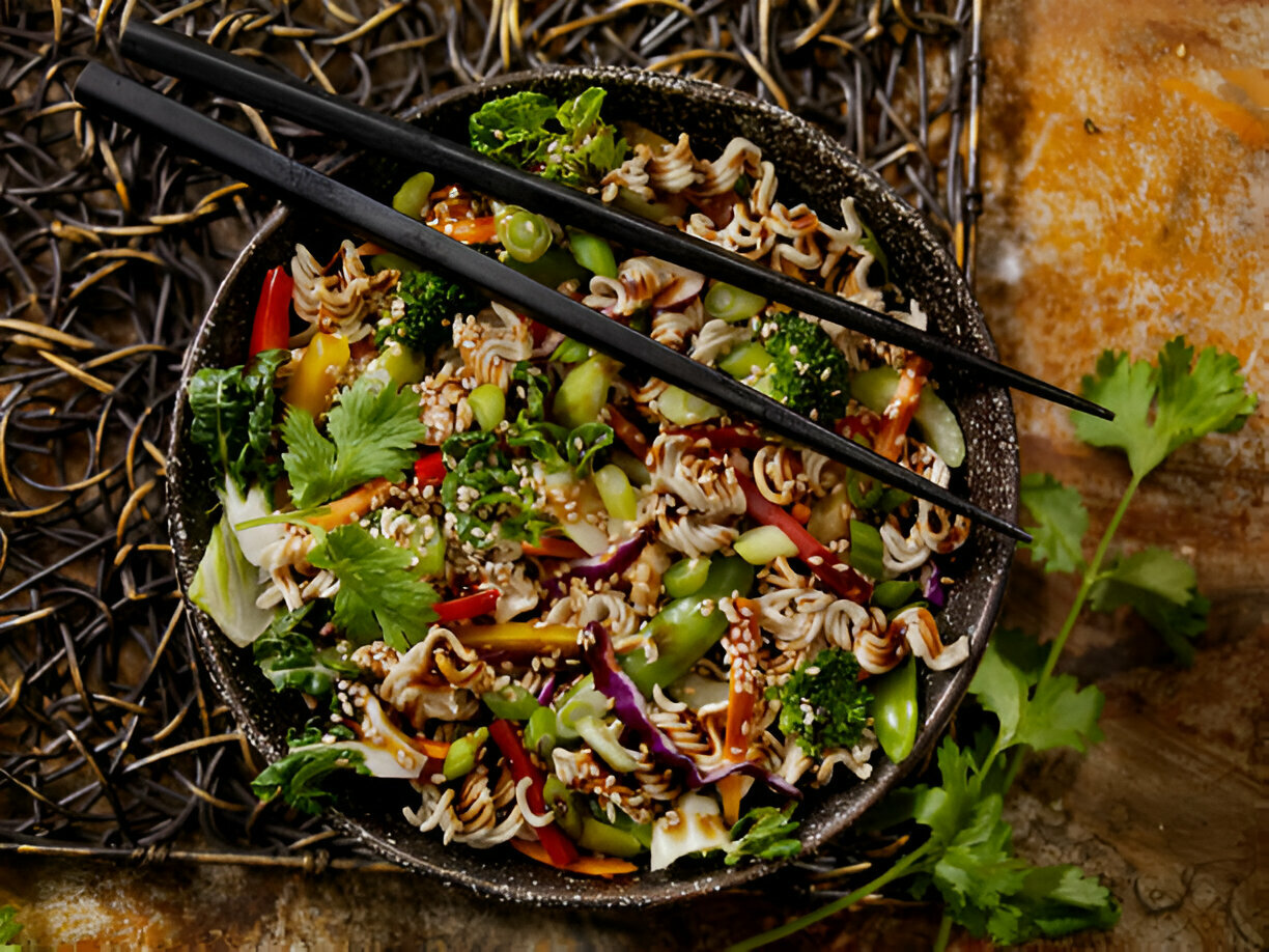 A bowl of colorful veggie noodle salad with sesame seeds and chopsticks, garnished with fresh cilantro.