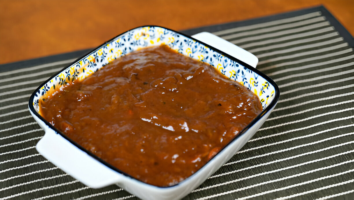 A dish of thick brown sauce in a decorated baking dish resting on a striped placemat.