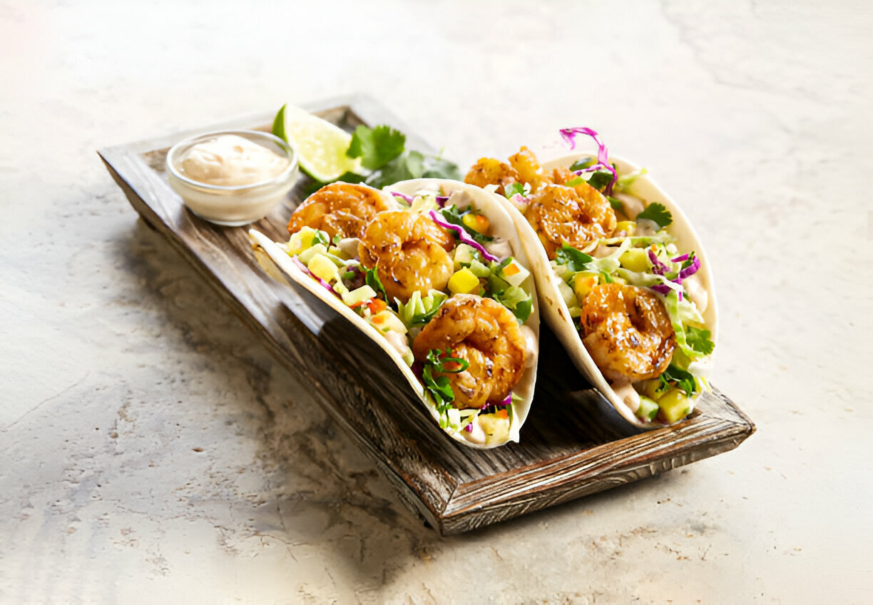 Three shrimp tacos with colorful toppings and a lime wedge on a wooden tray