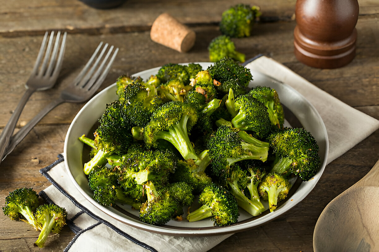 A plate of freshly cooked broccoli florets on a wooden table, alongside eating utensils.