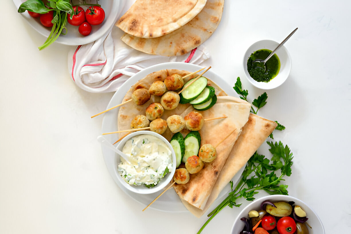 A plate of skewered meatballs served with pita bread, cucumber slices, and a creamy dipping sauce, alongside fresh herbs and tomatoes.