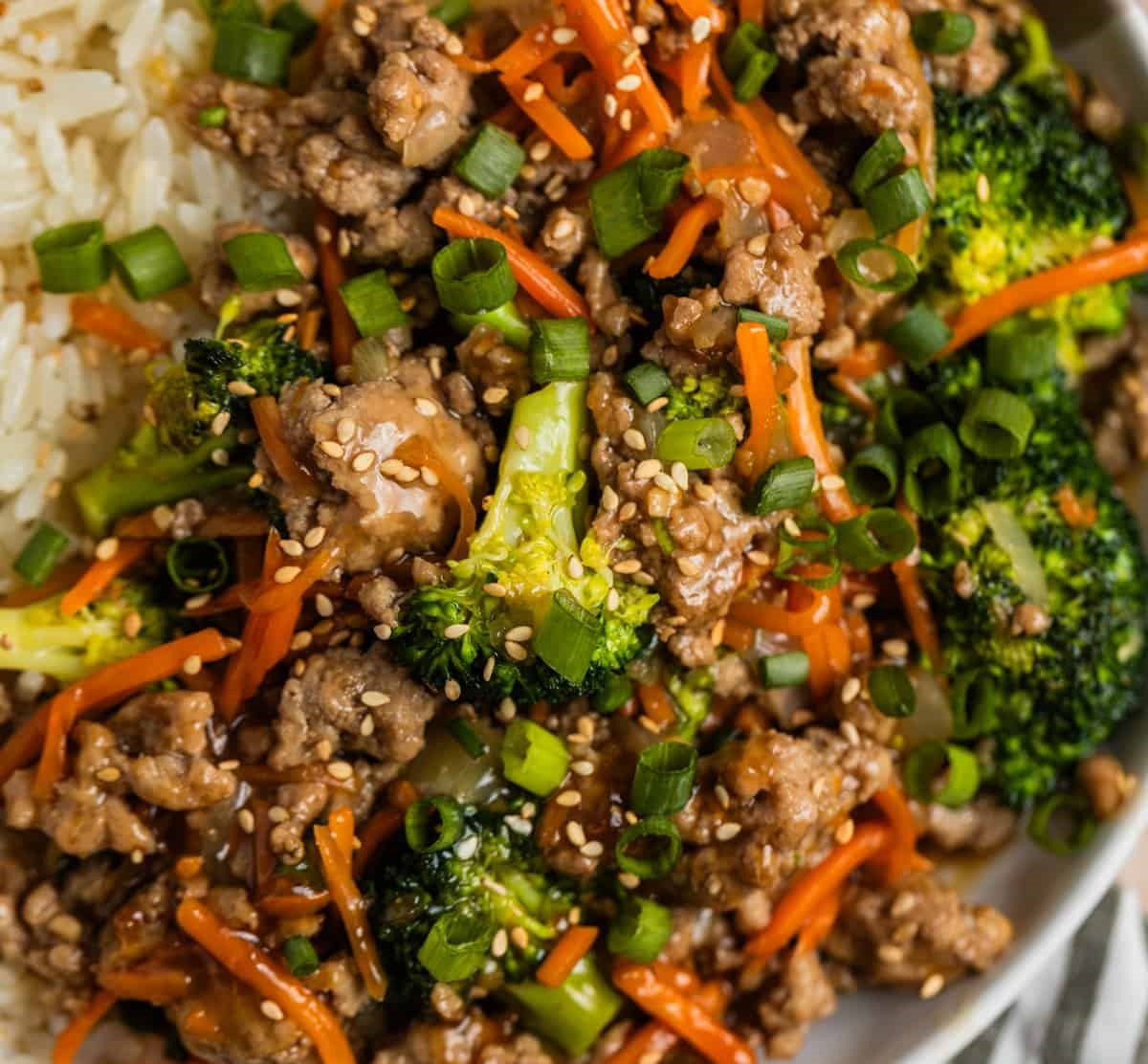 A delicious bowl of stir-fried beef and broccoli with sesame seeds, carrots, and green onions served alongside fluffy white rice.