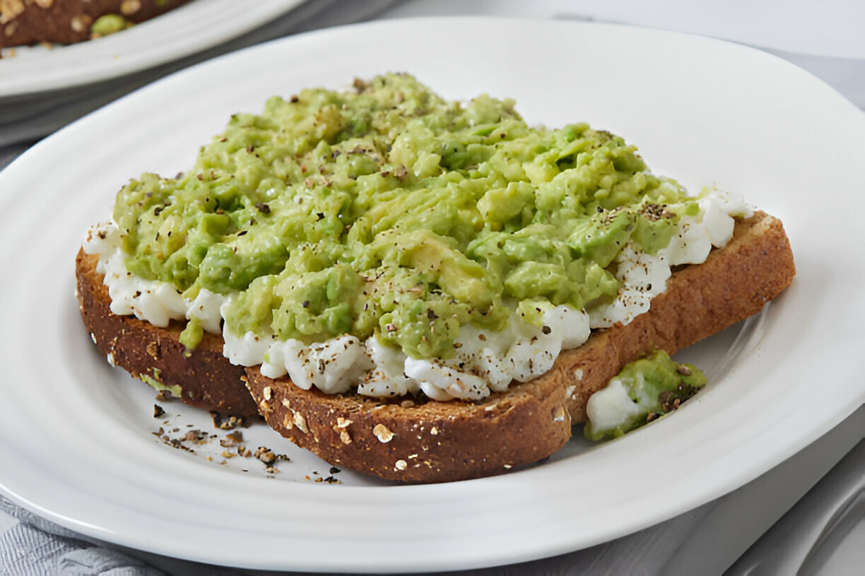 A plate of whole grain toast topped with mashed avocado and cottage cheese, sprinkled with black pepper.