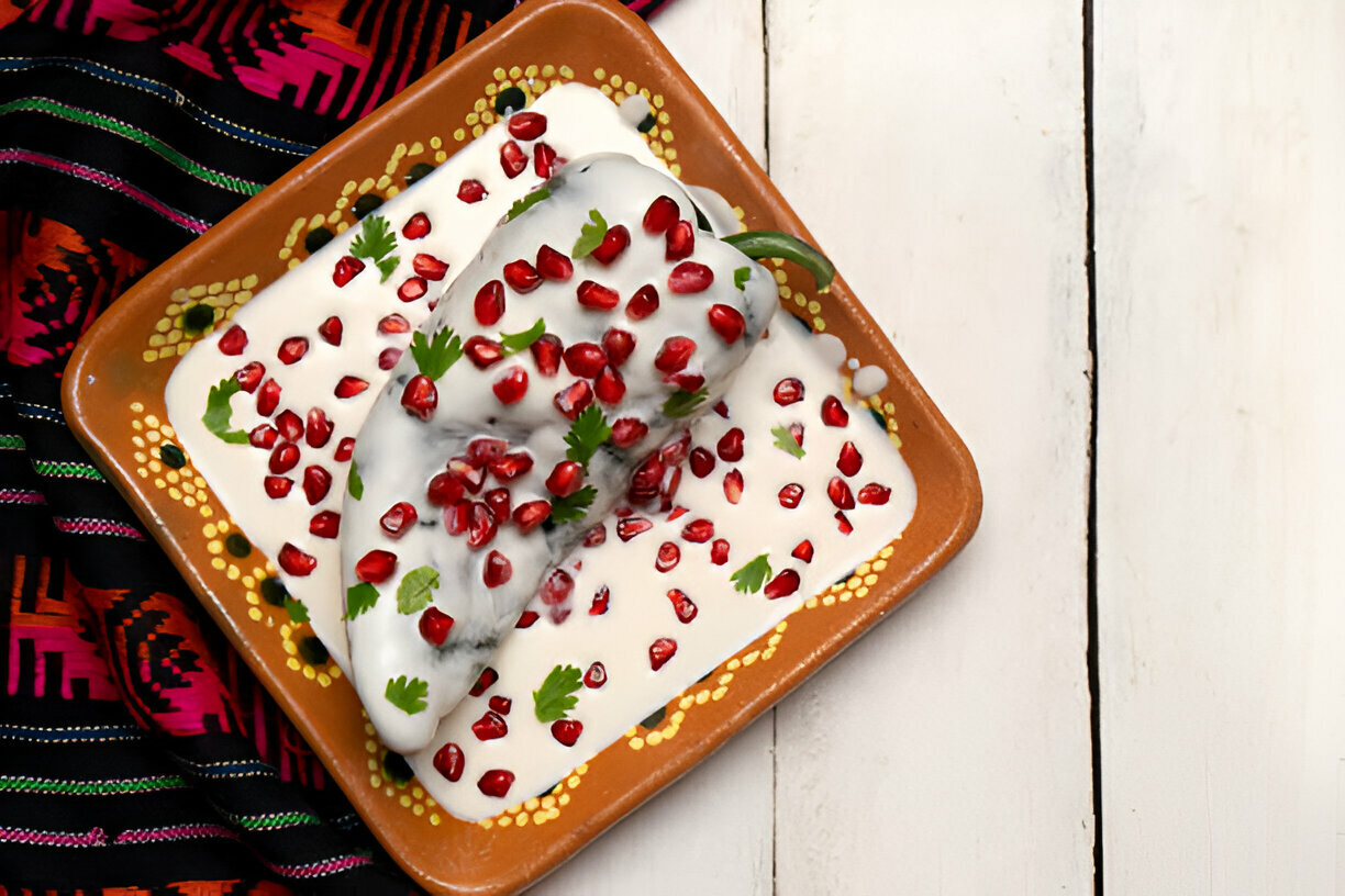 A dish with a green chili pepper covered in yogurt, garnished with pomegranate seeds and cilantro on a decorative plate, placed on a textured surface.