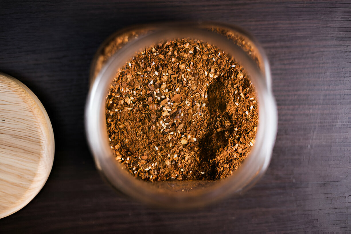 A close-up view of a jar containing brown spice or powder on a dark wooden surface, with a round wooden lid beside it.