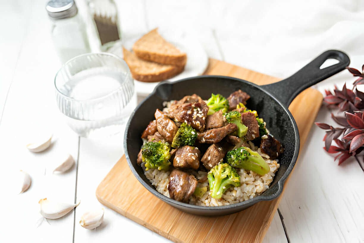 A skillet dish featuring pieces of meat and broccoli served over rice, with garlic cloves and a glass of water nearby.