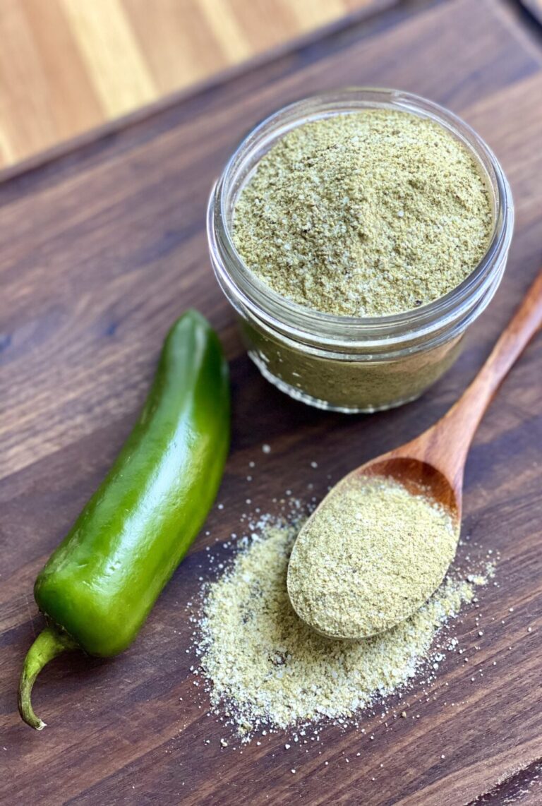 A glass jar filled with green spice powder next to a fresh green chili pepper and a wooden spoon on a wooden surface.