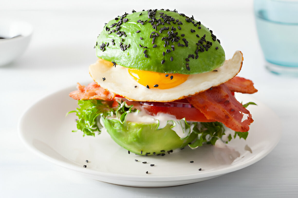 A gourmet dish featuring a stack of ingredients including bacon, lettuce, tomato, a fried egg, and a halved avocado topped with black sesame seeds on a white plate.