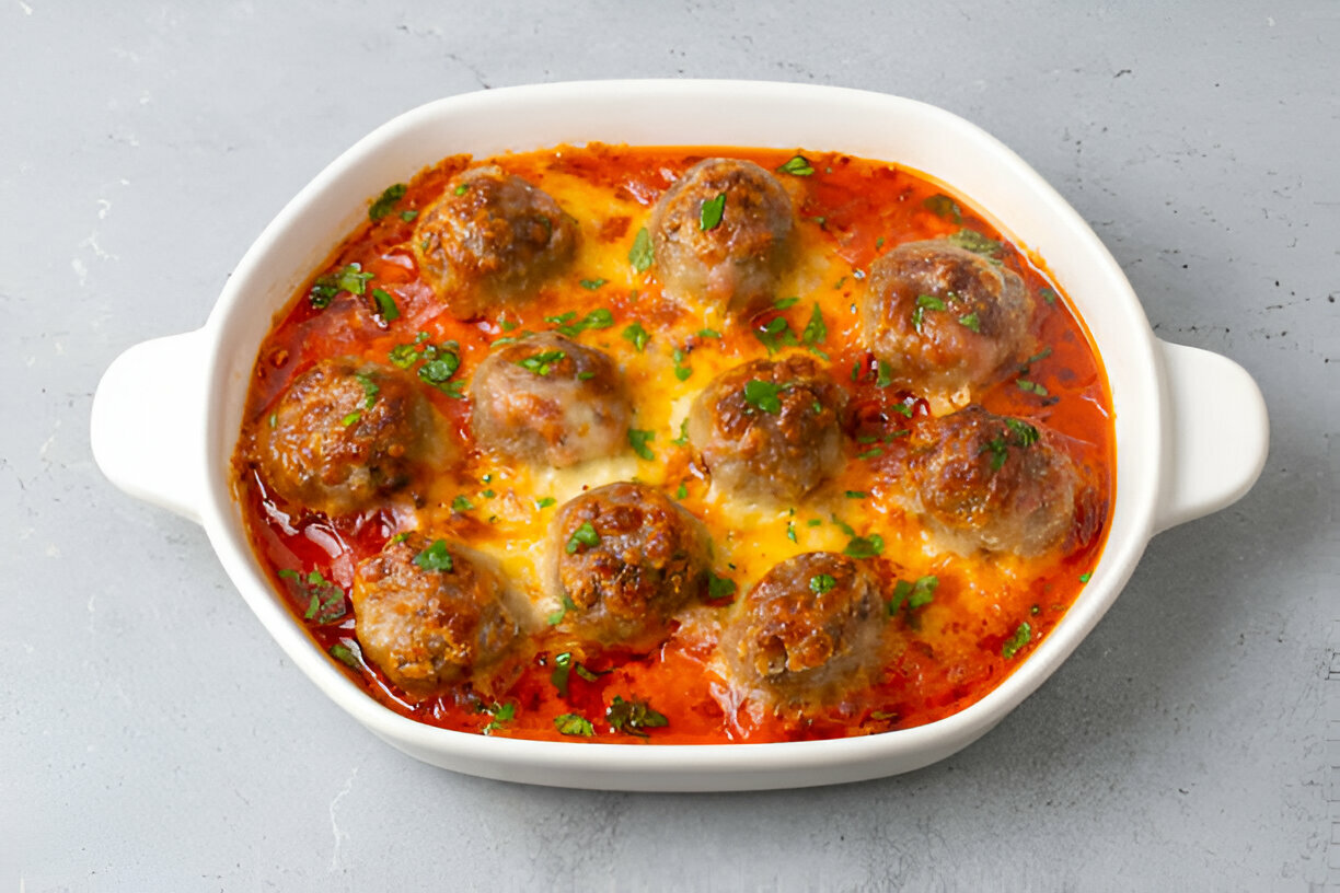 A dish of meatballs in tomato sauce topped with melted cheese and parsley, served in a white baking dish