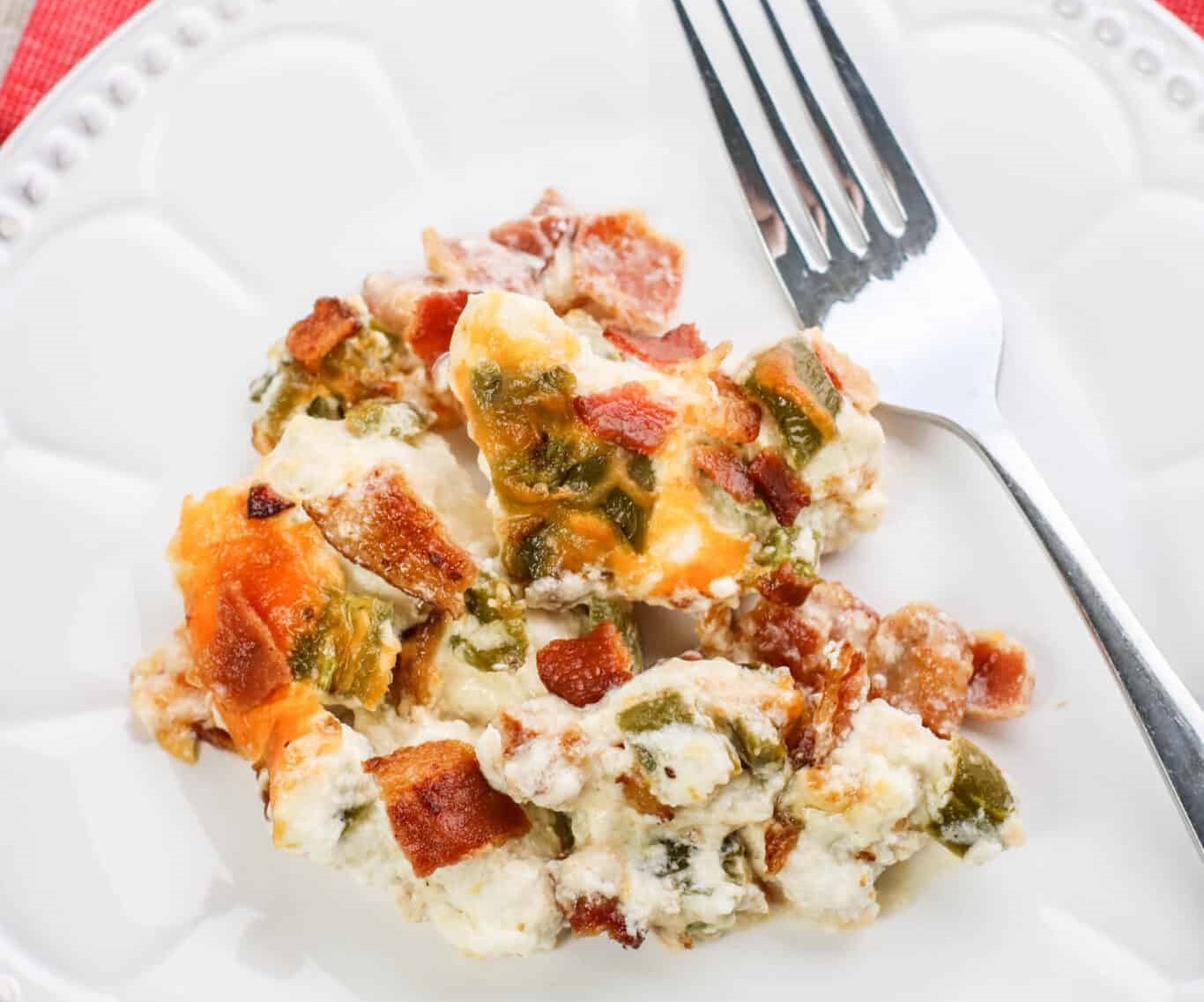 A serving of cheesy casserole with bacon and green peppers on a white plate, alongside a fork.