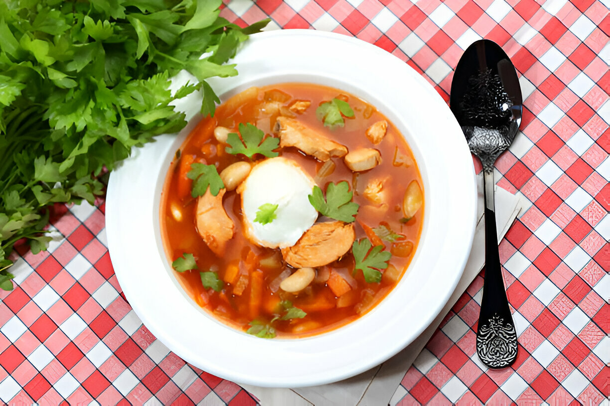 Bowl of chicken soup garnished with fresh cilantro on a red and white checkered tablecloth with a spoon on the side.