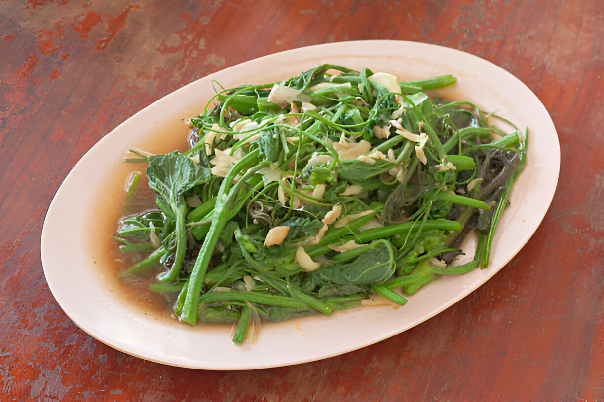 A platter of sautéed green vegetables garnished with sliced garlic on a wooden table.