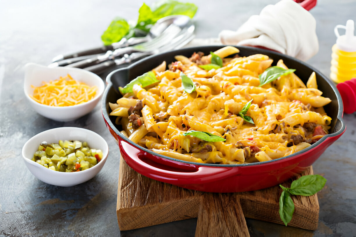 A delicious serving of cheesy pasta with ground beef, garnished with fresh basil, served in a red pan on a wooden cutting board.