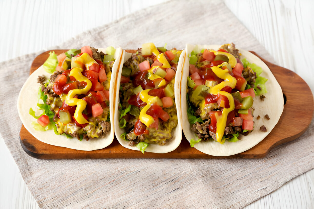 Three tacos topped with lettuce, diced tomatoes, pickles, mustard, and ketchup on a wooden serving board