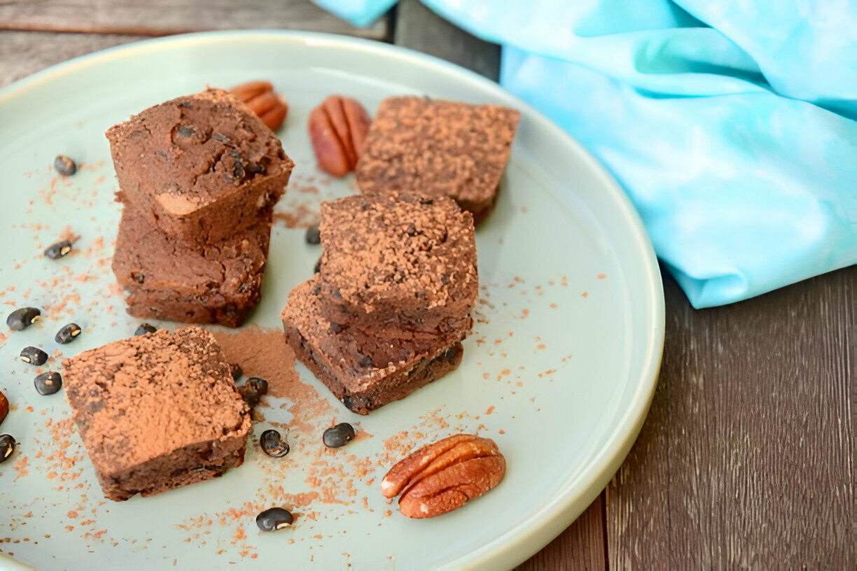 A stack of chocolate brownies sprinkled with cocoa powder on a light green plate, accompanied by pecans and cocoa nibs.