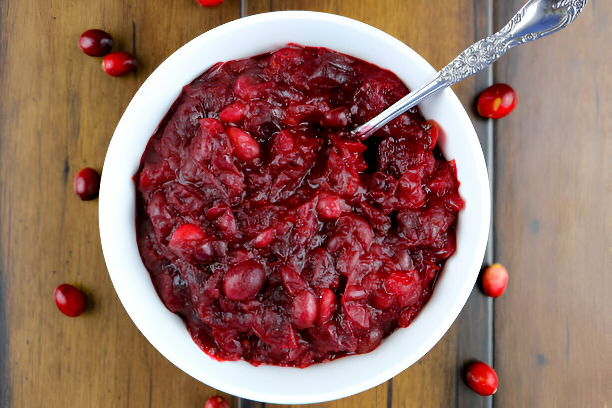 A bowl of homemade cranberry sauce placed on a wooden surface, surrounded by scattered cranberries.