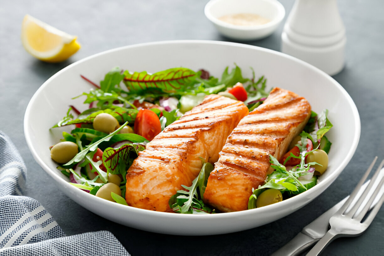 A bowl of grilled salmon on a bed of mixed greens, with cherry tomatoes and olives, garnished with lemon.