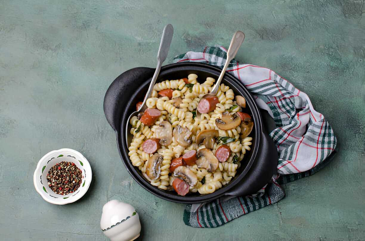 A serving of pasta with sausage, mushrooms, and herbs in a black bowl with two serving spoons.