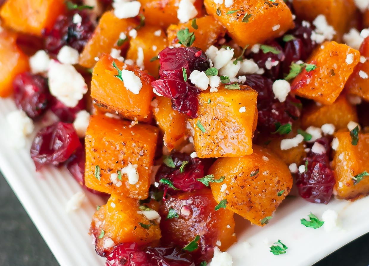 A close-up view of roasted butternut squash cubes mixed with dried cranberries and topped with crumbled feta cheese and fresh herbs.