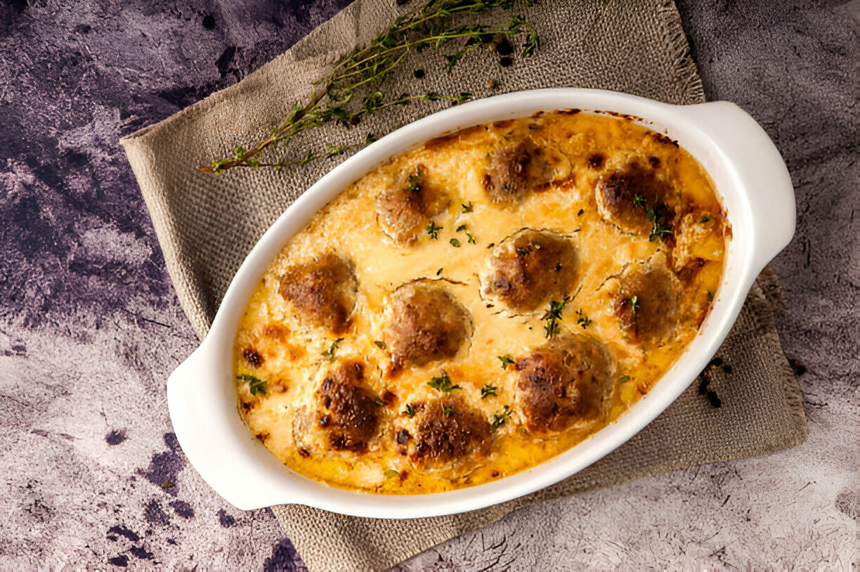 A baked casserole dish filled with golden meatballs in a creamy sauce, garnished with fresh herbs.