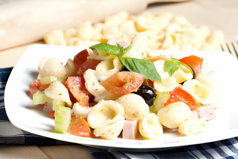 A close-up of a plate of pasta salad featuring tortellini, tomatoes, celery, black olives, and basil leaves.