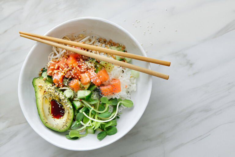 A vibrant bowl of sushi ingredients, including sliced avocado, diced salmon, cucumber, sprouts, and rice, garnished with sesame seeds and sauce, accompanied by chopsticks.