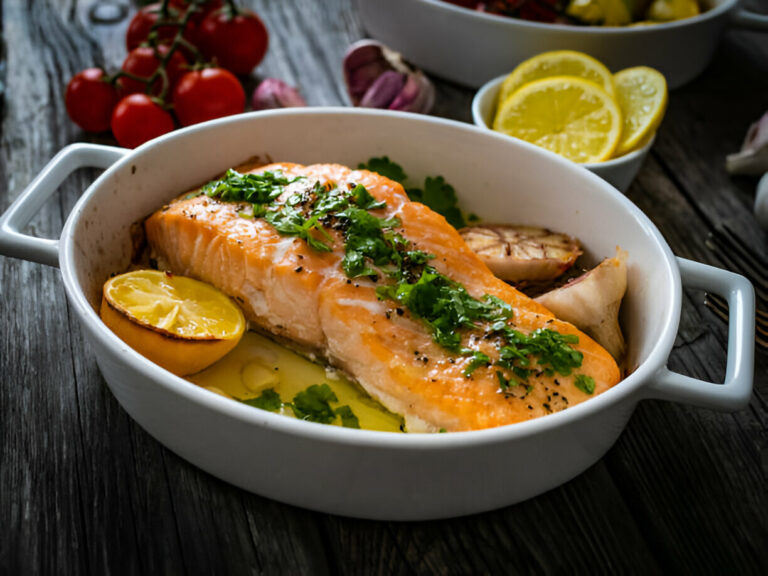 A serving dish with a cooked salmon fillet topped with herbs and accompanied by lemon slices.