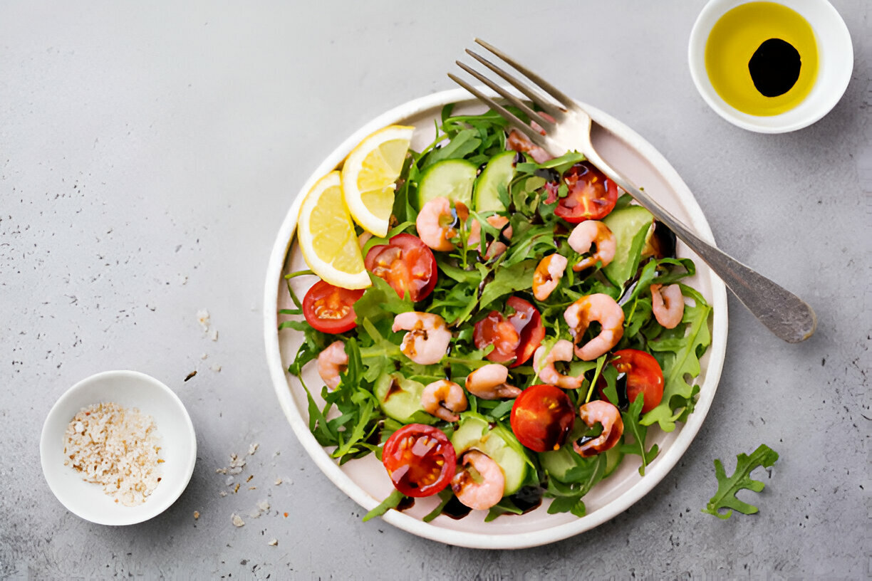 A vibrant salad with shrimp, mixed greens, cucumber slices, halved cherry tomatoes, and lemon wedges on a white plate, accompanied by olive oil and salt in small bowls.