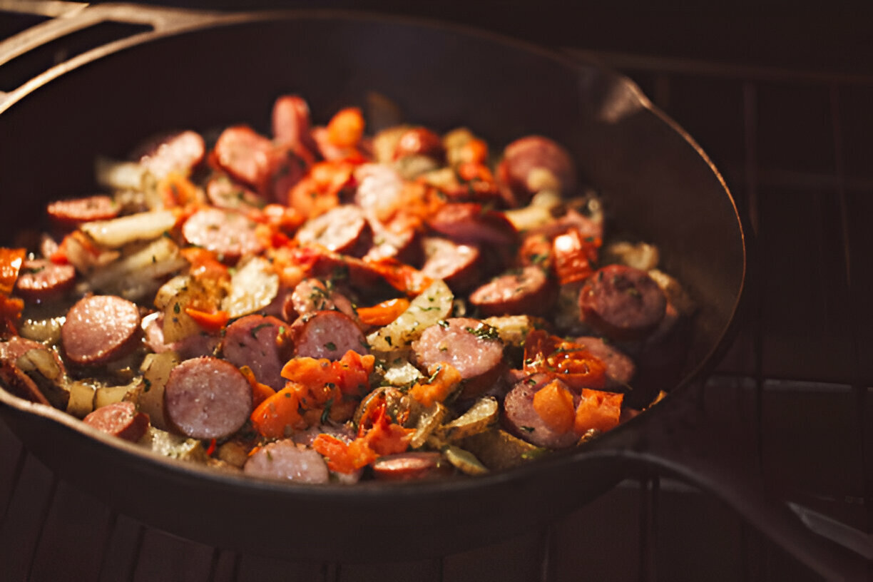 A cast iron skillet filled with sautéed sausages, bell peppers, onions, and herbs cooking on a stovetop.