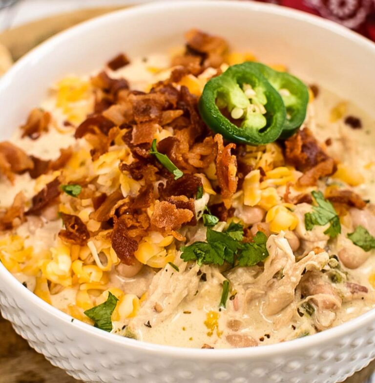 A close-up of a bowl of creamy white chicken chili topped with crispy bacon, shredded cheese, fresh cilantro, and sliced jalapeño.