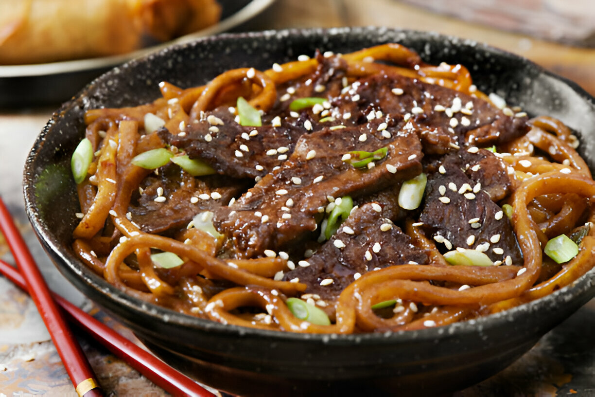 A delicious bowl of beef udon noodles garnished with sesame seeds and scallions, served alongside chopsticks.