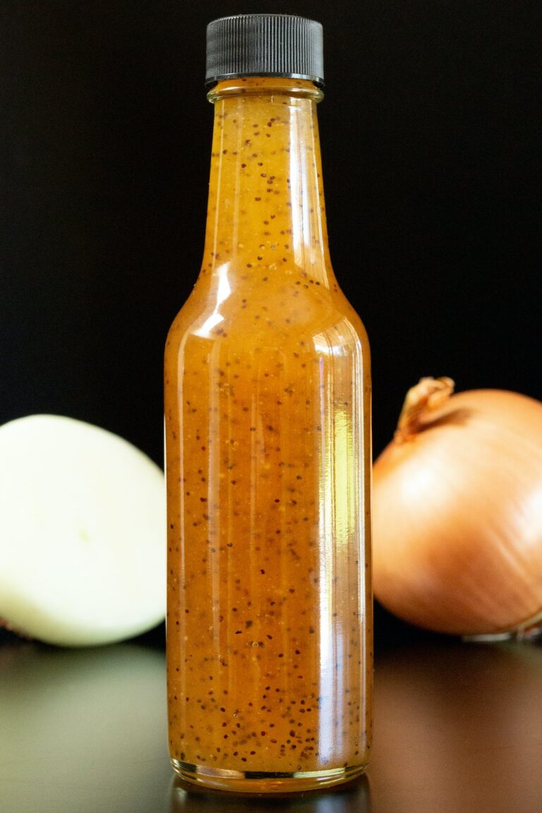 A bottle of sauce with a black cap, next to an onion and part of a white onion on a dark surface.