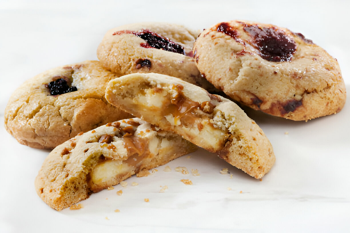 A close-up of several baked cookies, some with jelly on top and one cut in half revealing a gooey caramel and chocolate filling.