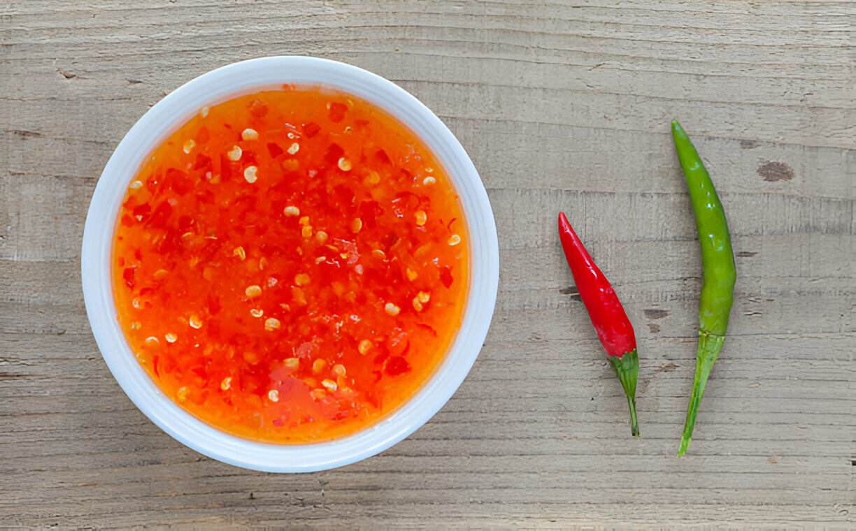 A small bowl filled with spicy chili sauce, surrounded by red and green chili peppers on a wooden surface.