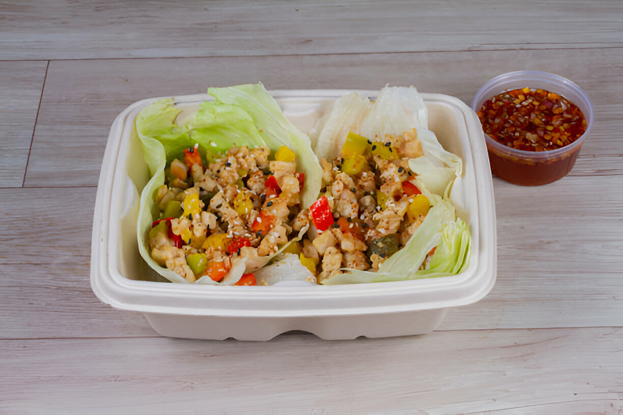 A takeout container with lettuce wraps filled with diced vegetables and protein, accompanied by a small container of sauce.