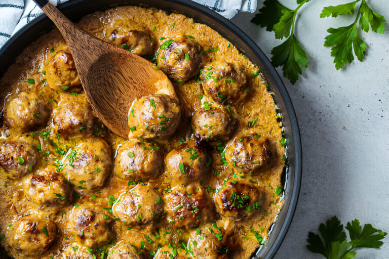 A skillet filled with meatballs covered in a creamy sauce, garnished with parsley and a wooden spoon resting on the side.