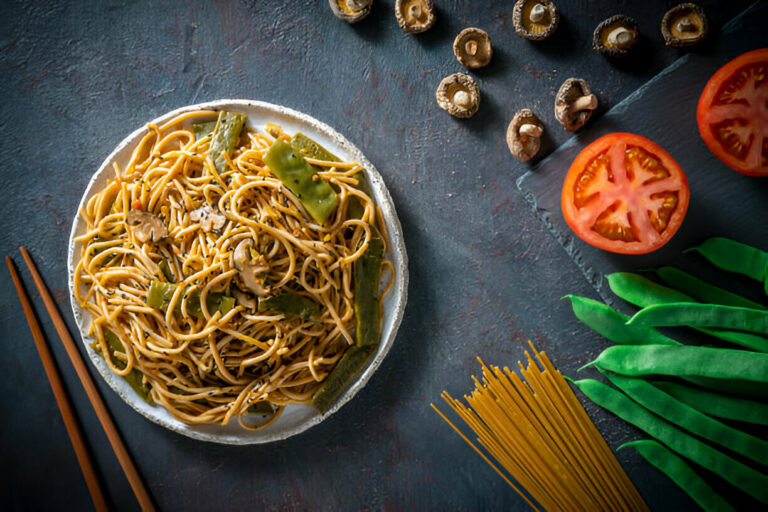 A plate of noodles with vegetables, surrounded by fresh tomatoes and green beans on a textured background.